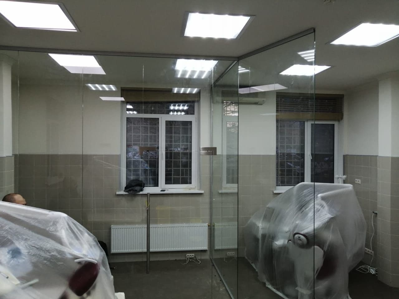 Кабинеты клиники за прозрачными полотнами: дверь из стекла с длинной ручкой-скобой Перегородки от производителя в стоматологии: стекло без стоек делит кабинеты, кресла ещё под плёнкой